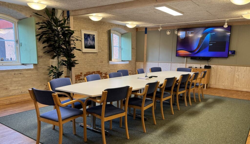 Logen - Ett konferensrum med ett långt vitt bord och 14 stolar. På väggen sitter en stor skärm.