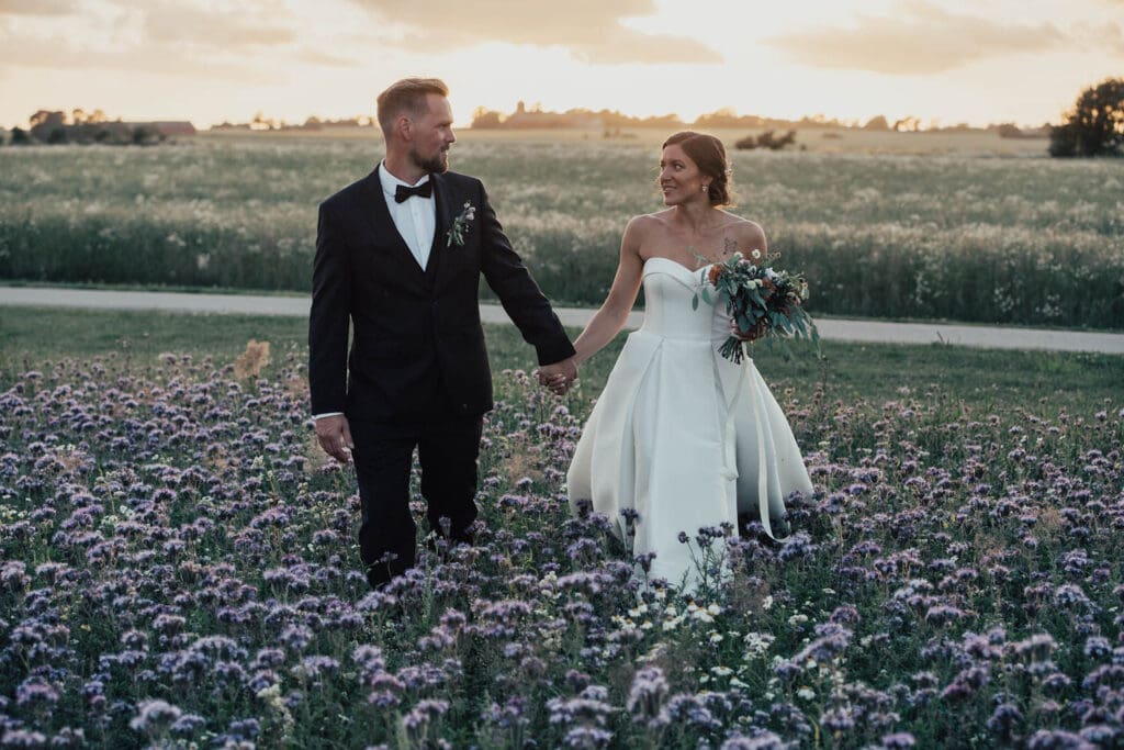 En man och en kvinna i bröllopsklädsel. mannen har svart kostym och kvinnan vit klänning. De går mot kameran i ett fält av lila blommor