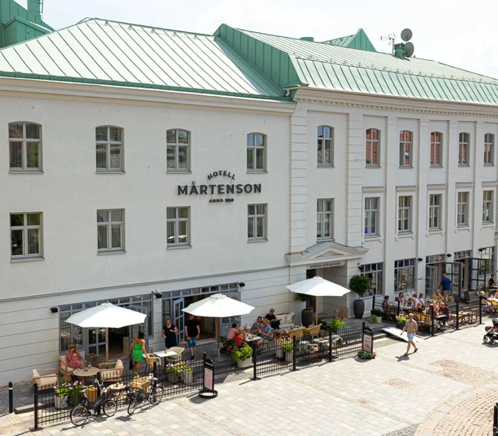 En vit större byggnad med grönt tak i en stad. Uteservering med parasoller utanför i solen 